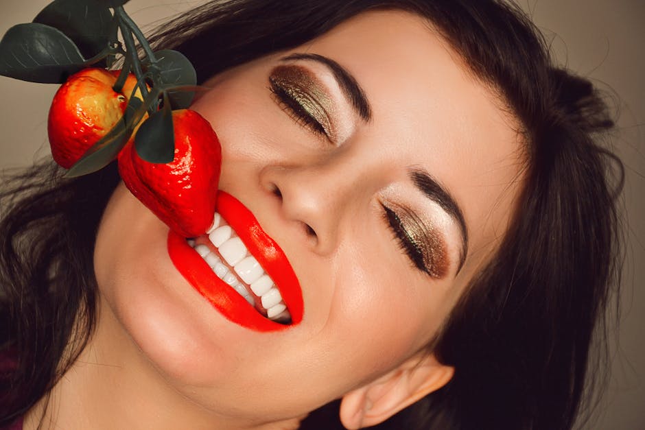 Close-up of a woman with vibrant makeup and red lips smiling next to artificial strawberries.